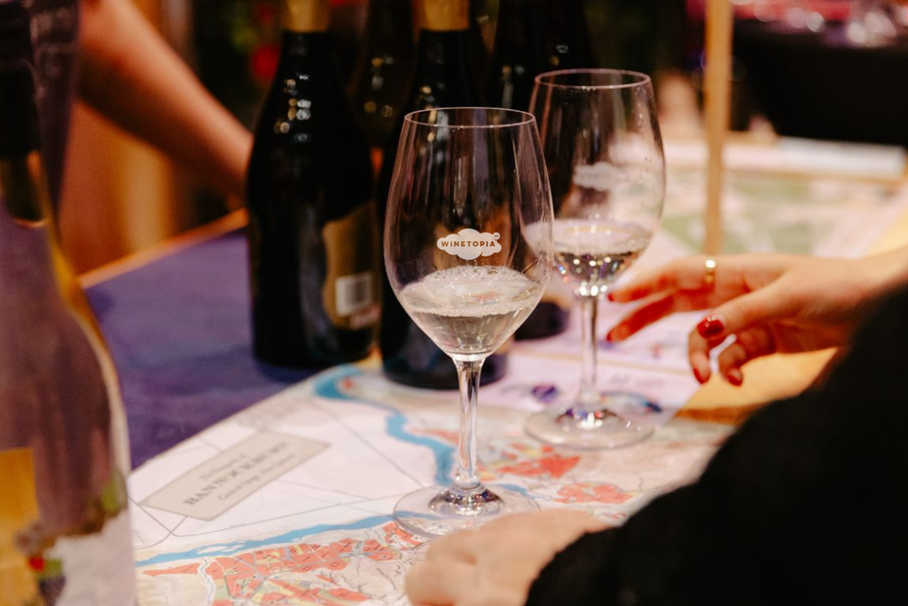 Two wine glasses on a table with bottles and a person's hand in the background